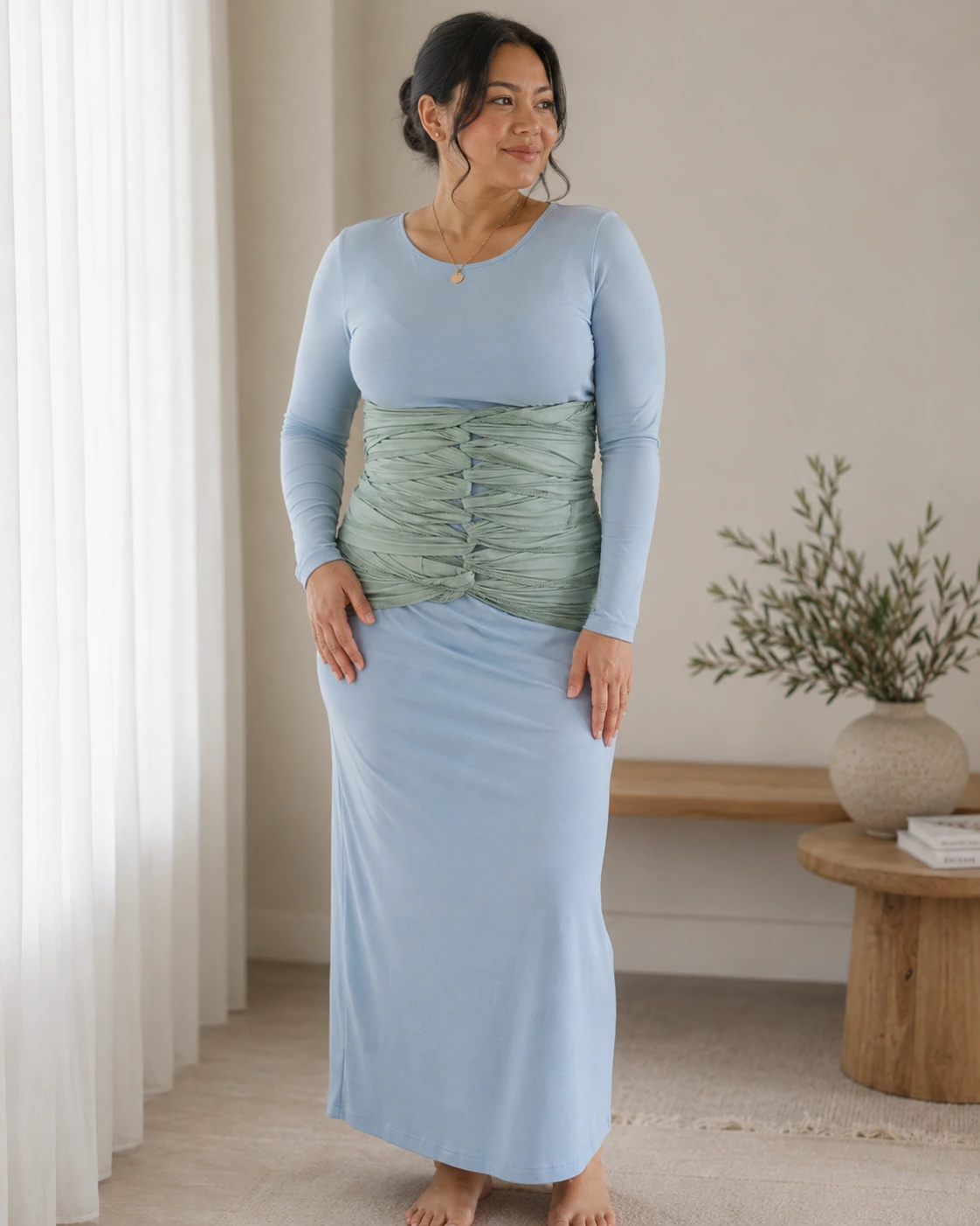 Postpartum mother standing in a soft blue dress with a sage belly wrap in a full-length recovery portrait
